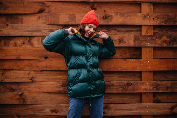 Fashionable girl in great mood posing on wooden background. Woman in baggy jeans, emerald coat and red hat smiling.
