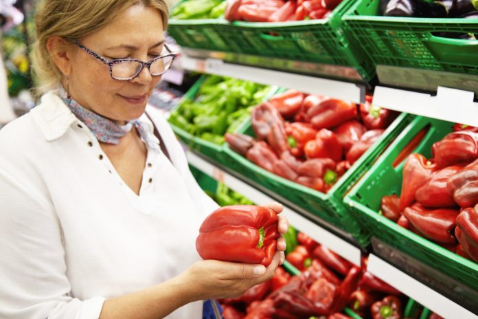 Good-looking senior woman wearing glasses holding red pepper, examining it with care, smiling while grocery shopping in supermarket, standing in front of boxes filled with fresh fruits and vegetables