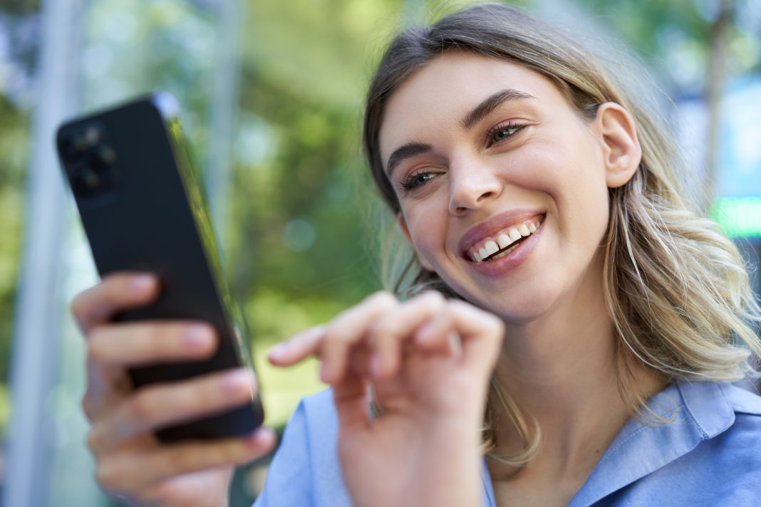Beautiful young woman sitting outdoors on street, smiling at smartphone, buying online, order on mobile phone