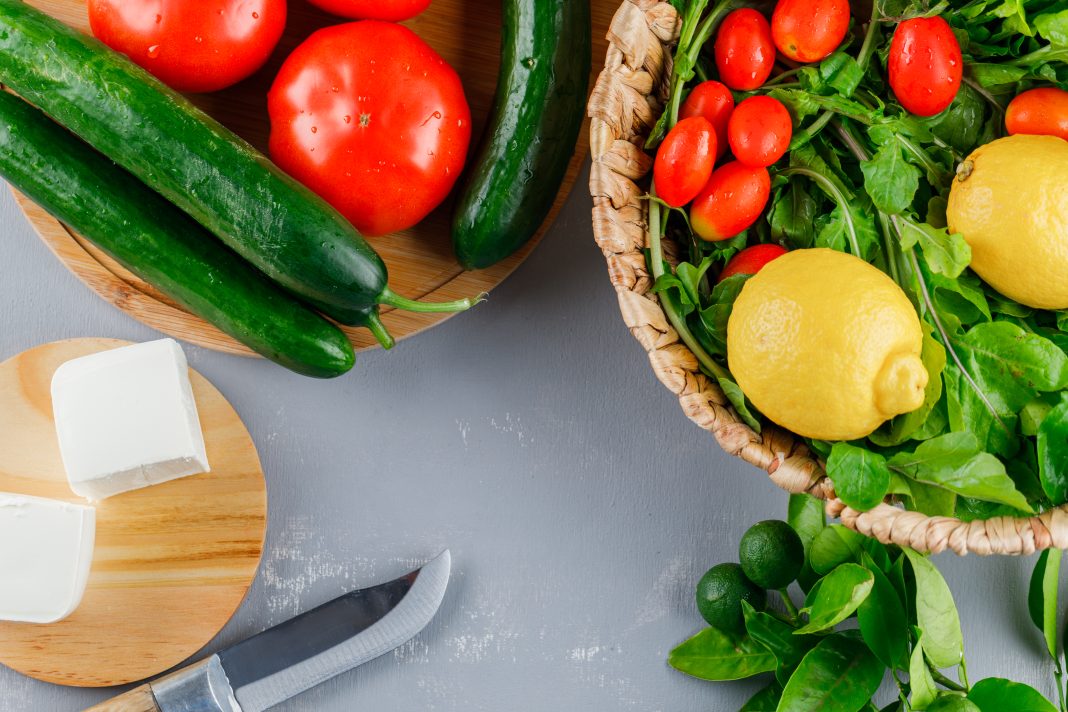 Set of knife, lemon, cucumber, cheese, greens and tomatoes in a cutting board on a gray background. top view.